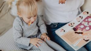Cute toddler with ponytails enjoying a snack next to a book and parent. Cozy indoor scene.